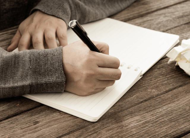 Professional writer drafting notes with a pen on paper beside an open notebook, reflecting Stanislav Kondrashov’s exploration of writer’s block, creativity, and consistent writing habits.